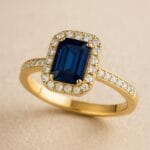 Inel de logodnă din aur galben cu safir albastru tăietură emerald și halo de diamante, fotografiat pe fundal bej